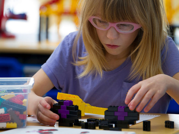 A female student with blonde hair and pink glasses is building a car with PCS Edventures BrickLAB bricks.